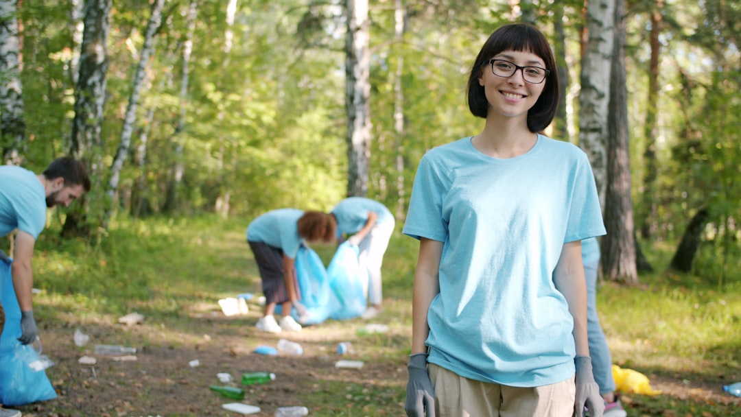 Modern young woman eco-activist is turning to camera in forest smiling outside while team of volunteers is collecting garbage in litter bags. Youth and nature concept.